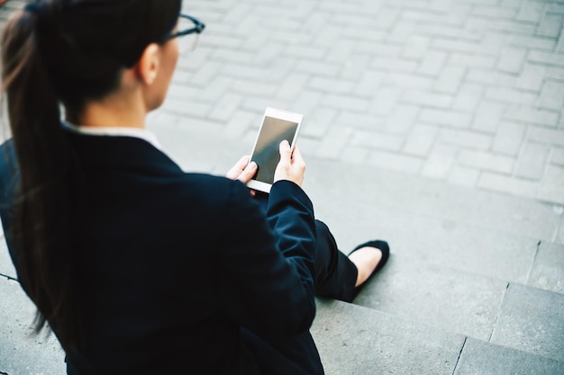 Femme d'affaires heureuse Femme souriante et joyeuse dans un costume d'affaires et des lunettes assis sur des marches en béton gris tient le téléphone dans ses mains