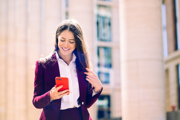 Femme d'affaires heureux lire les bonnes nouvelles dans son smartphone large sourire. Cadre travaillant avec un téléphone portable dans la rue avec des immeubles de bureaux en arrière-plan