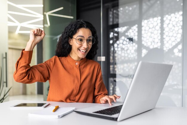 Femme d'affaires hispanique célébrant la réussite de la victoire employée avec des cheveux bouclés à l'intérieur de la lecture de bureau