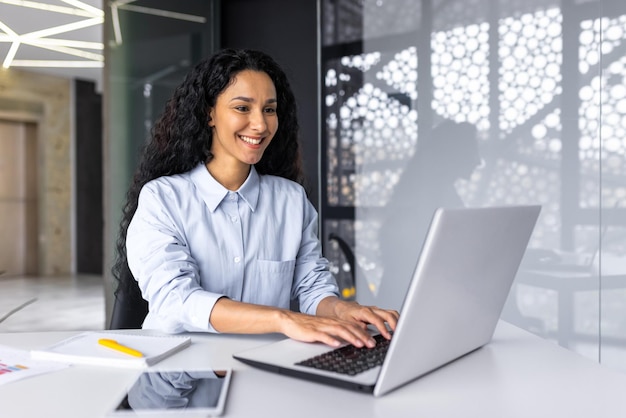 Femme d'affaires hispanique heureuse et souriante tapant sur un employé de bureau d'ordinateur portable avec des cheveux bouclés et des lunettes