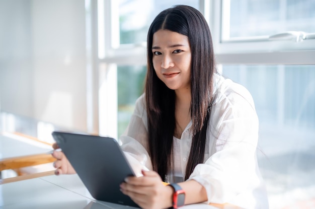 Femme d'affaires indépendante asiatique portant une montre intelligente parlant en utilisant un travail occasionnel avec une tablette pour naviguer sur le chat et les blogs sur Internet et un smartphonenotebook dans un café