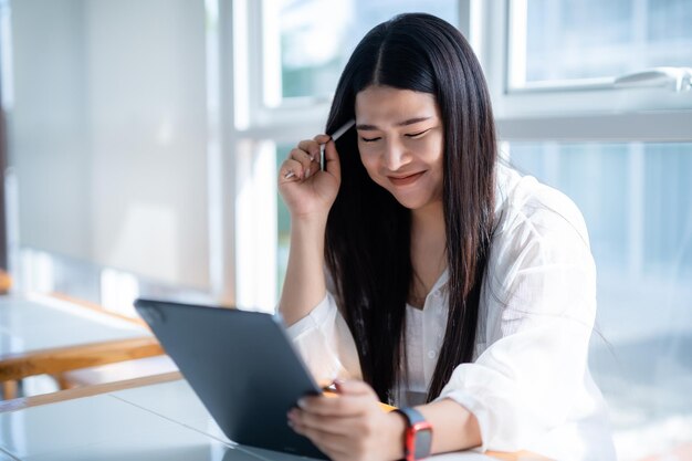 Femme d'affaires indépendante asiatique portant une montre intelligente parlant en utilisant un travail occasionnel avec une tablette pour naviguer sur le chat et les blogs sur Internet et un smartphonenotebook dans un café