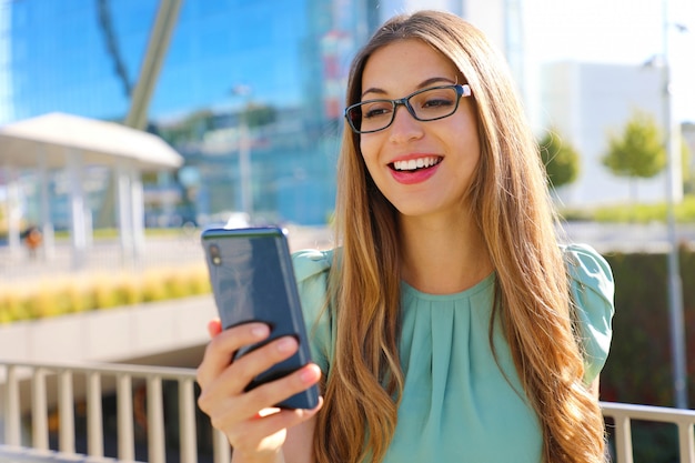 Femme d'affaires intelligente souriante avec téléphone portable dans la rue avec des immeubles de bureaux en arrière-plan.