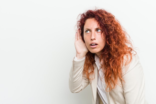 Femme d'affaires jeune rousse naturelle isolée contre le mur blanc essayant d'écouter un potin.