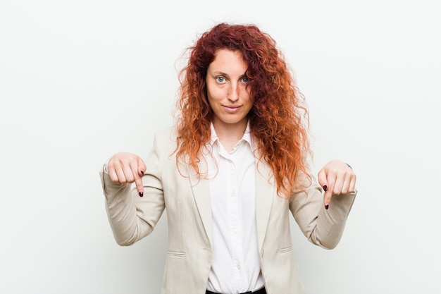 Femme d'affaires jeune rousse naturelle isolée contre le mur blanc pointe vers le bas avec les doigts, sentiment positif.