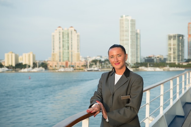 Femme d'affaires ou jolie fille heureuse avec un visage souriant en veste grise, a une manucure rouge sur le pont du navire avec vue sur la mer, l'eau de mer avec un bateau à voile sur fond de ciel nuageux, les voyages et le tourisme