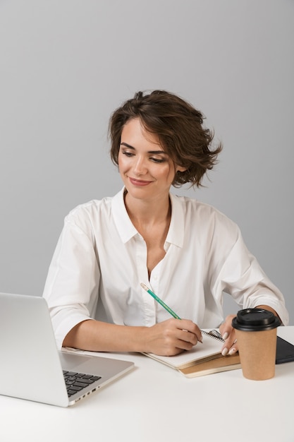 Femme d'affaires joyeuse heureuse posant isolé sur mur gris assis à la table à l'aide d'un ordinateur portable