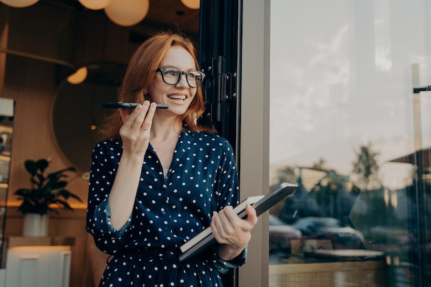 Une femme d'affaires joyeuse et joyeuse au gingembre indépendante avec un ordinateur portable tient un téléphone portable et parle sur haut-parleur avec un ami ou un client, utilise un assistant virtuel ou enregistre un message audio sur un smartphone à l'extérieur