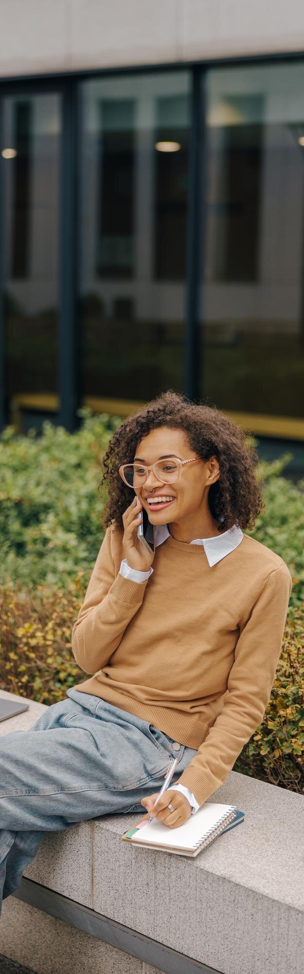 Une femme d'affaires joyeuse parle au téléphone et prend des notes alors qu'elle est assise sur l'arrière-plan du bâtiment