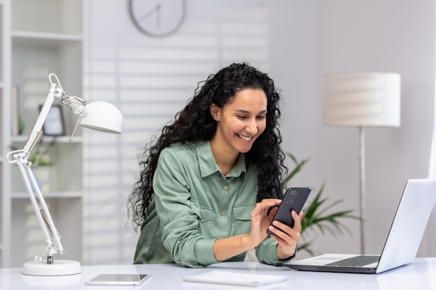 Femme d'affaires joyeuse sur son lieu de travail en utilisant son téléphone latino-américaine heureuse en utilisant l'application sur son smartphone en naviguant