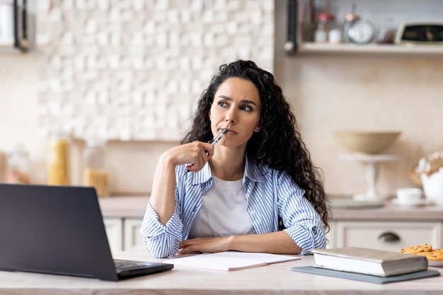 Femme d'affaires latine sérieuse planifiant sa journée assise devant un ordinateur portable dans la cuisine pensant et