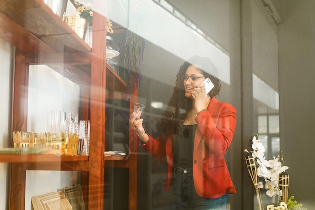 Femme d'affaires latino-américaine tenant un verre d'une étagère d'articles et passant un appel téléphonique avec un mobile blanc