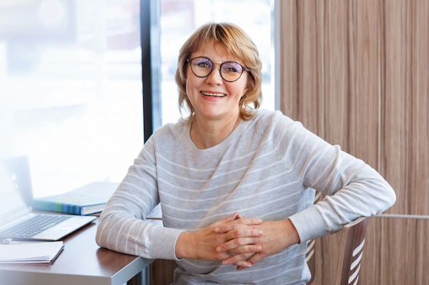 Femme d'affaires sur le lieu de travail au bureau. Une femme d'âge moyen travaille sur un ordinateur portable dans un café. Elle porte des lunettes et sourit.