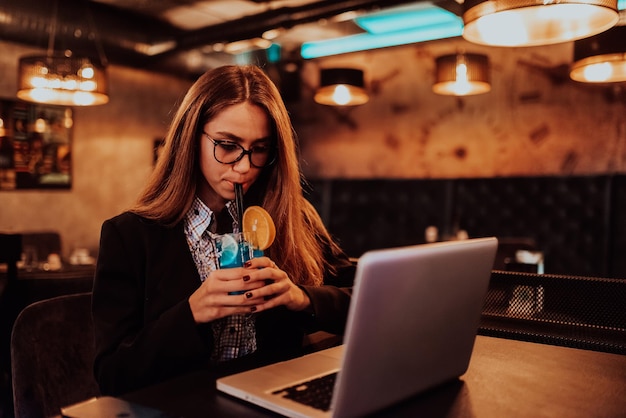 Femme d'affaires à lunettes assise dans un café moderne et travaillant sur son ordinateur portable tout en buvant un cocktail Mise au point sélective