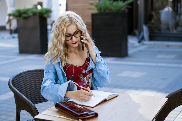 Femme d'affaires avec des lunettes est assise dans un café dans la rue, écrit dans un ordinateur portable et parle au téléphone