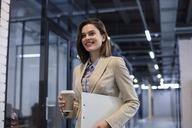 Femme d'affaires marchant le long du couloir du bureau avec des documents et un gobelet en papier.