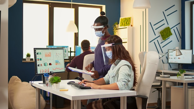 Femme d'affaires avec masque de protection expliquant à un collègue africain les graphiques du presse-papiers pointant sur le bureau de l'ordinateur, assise au bureau dans un nouveau bureau normal. Equipe multiethnique respectant la distanciation sociale.