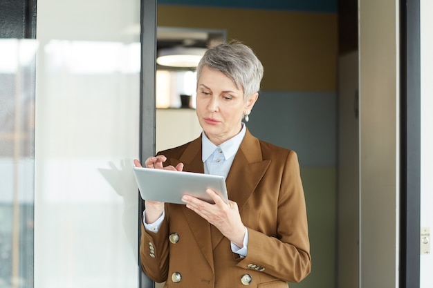 Femme d'affaires mature aux cheveux gris à l'aide de tablette numérique près de l'entrée du bureau