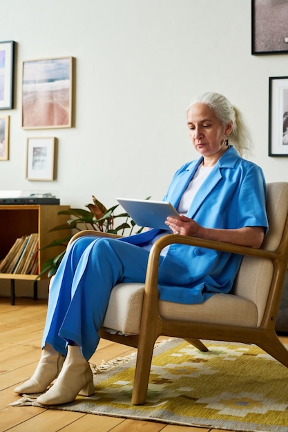 Femme d'affaires mature aux longs cheveux gris à l'aide d'une tablette assise dans un fauteuil