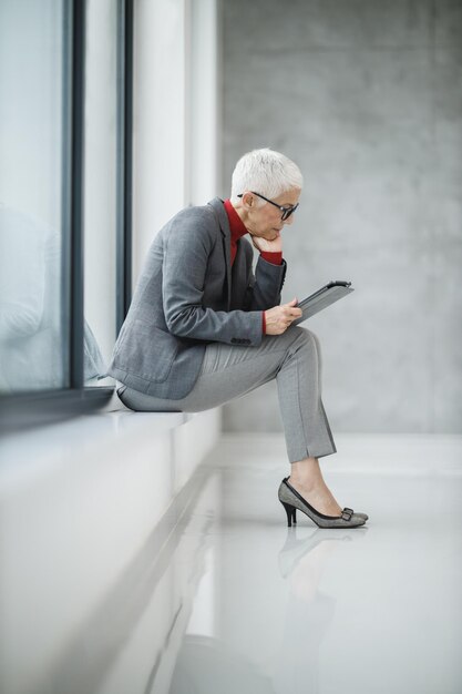 Une femme d'affaires mature confiante travaillant sur une tablette numérique tout en étant assise près de la fenêtre dans un bureau moderne.