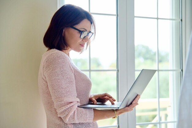 Femme d'affaires mature dans des verres avec un ordinateur portable dans les mains près de la fenêtre