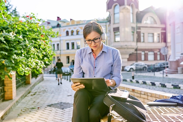 Femme d'affaires mature sérieuse dans des écouteurs à lunettes avec tablette numérique dans la rue de la ville