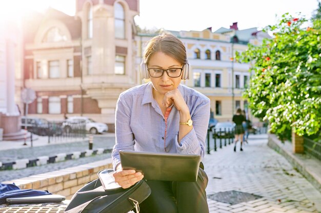 Femme d'affaires mature sérieuse dans des écouteurs à lunettes avec tablette numérique dans la rue de la ville