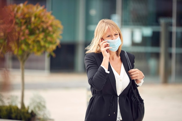 Une femme d'affaires mature avec un téléphone portable portant un masque facial EPI marche dans la rue pendant la pandémie de santé