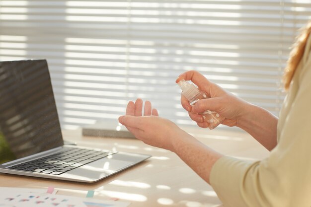 Femme d'affaires méconnaissable pulvérisant les mains avec un désinfectant sur le lieu de travail éclairé par la lumière du soleil