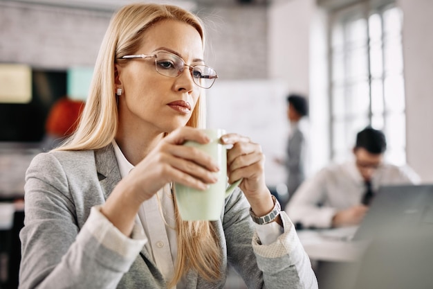 Femme d'affaires mécontente de boire du thé et de penser à quelque chose tout en travaillant au bureau Il y a des gens en arrière-plan