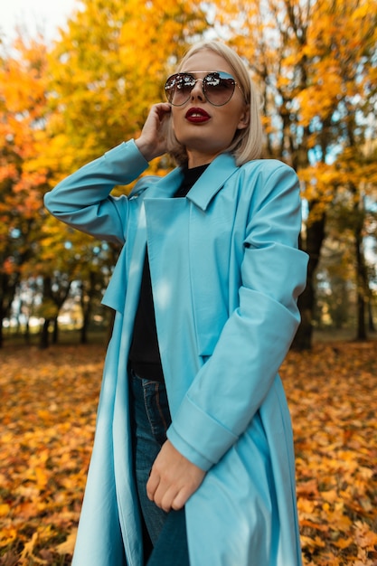 Femme d'affaires de mode avec des lunettes de soleil dans un élégant manteau bleu à pied dans un parc en automne avec des feuilles d'automne jaunes