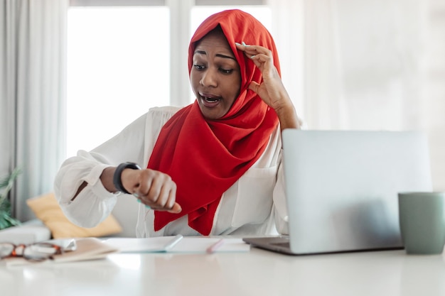 Femme d'affaires musulmane afro-américaine assise au bureau devant un ordinateur portable regardant la montre et touchant la tête