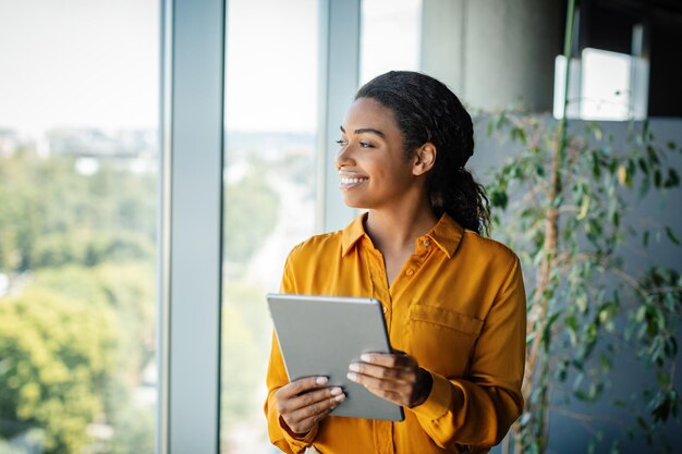 Femme d'affaires noire excitée debout dans une salle de bureau de coworking moderne regardant à travers l'espace de copie de fenêtre