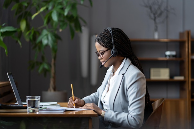 Une femme d'affaires noire joyeuse dans un casque étudie en ligne des notes d'écriture en regardant un cours de webinaire sur un ordinateur portable