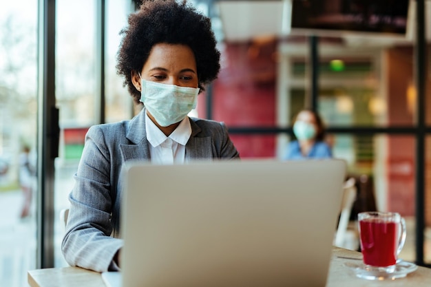 Femme d'affaires noire portant un masque de protection sur son visage tout en travaillant sur un ordinateur portable dans un café