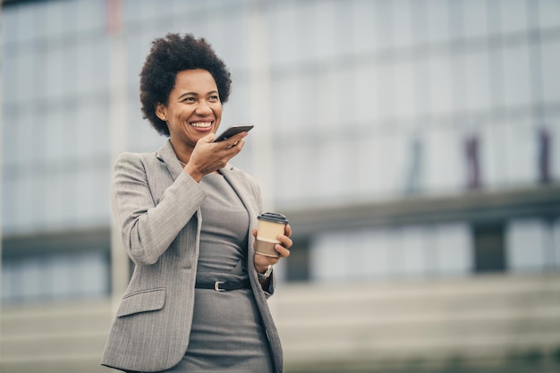 Une femme d'affaires noire prospère parlant un message vocal sur un téléphone intelligent tout en prenant une pause-café devant un bâtiment d'entreprise.