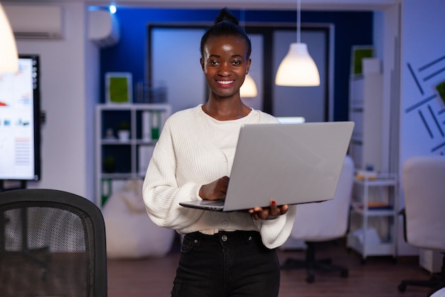 Femme d'affaires noire regardant la caméra souriante tenant un ordinateur portable debout près du bureau dans une entreprise en démarrage tard dans la nuit