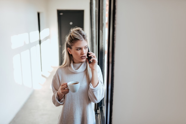 Femme d&#39;affaires occasionnelle confiante, parlant au téléphone. Tenant une tasse de café et regardant par la fenêtre.