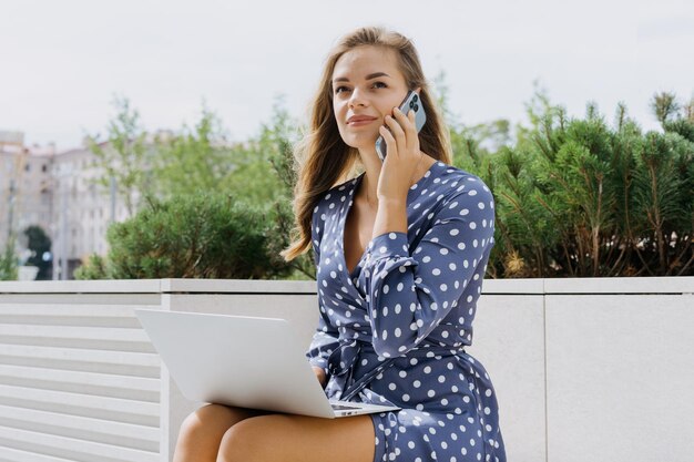 Une femme d'affaires avec un ordinateur portable travaille assise sur un banc et parle au téléphone