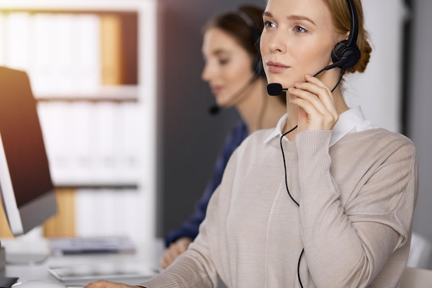 Femme d'affaires parlant par casque alors qu'il était assis dans un bureau ensoleillé Centre d'appels et divers groupes de personnes dans les affaires