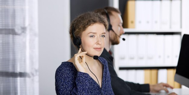 Femme d'affaires parlant par casque tout en étant assis avec un collègue à barbe rouge dans un bureau moderne. Télémarketing et service à la clientèle. Groupe de personnes diverses dans le centre d'appels.