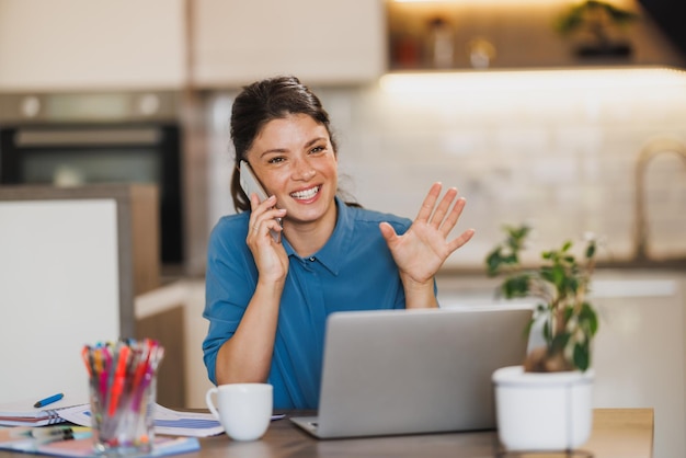 Femme d'affaires parlant sur un smartphone tout en travaillant sur un ordinateur portable depuis son bureau à domicile.