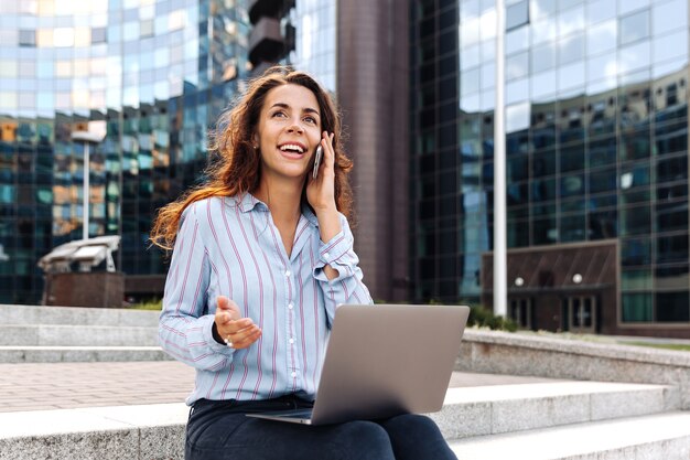 Femme d'affaires parlant sur un téléphone portable et travaillant sur un ordinateur portable dans la rue