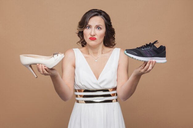 Femme d'affaires à la pensée douteuse en robe blanche debout, tenant des talons hauts élégants et des baskets confortables et ne sait pas laquelle choisir. Studio shot, intérieur, isolé sur fond marron clair