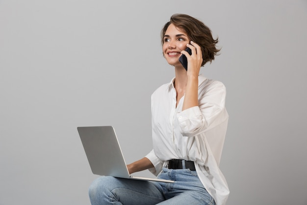 Femme d'affaires posant isolé sur mur gris assis sur un tabouret à l'aide d'un ordinateur portable parler par téléphone