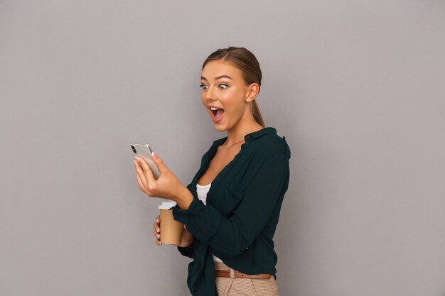 Femme d'affaires posant isolée sur fond de mur gris, boire du café à l'aide d'un téléphone portable.