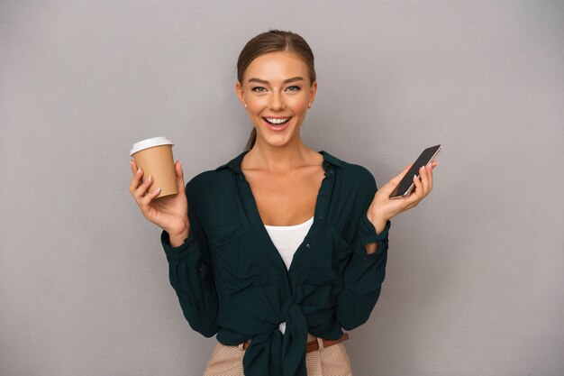 Femme d'affaires posant isolée sur fond de mur gris, boire du café à l'aide d'un téléphone portable.