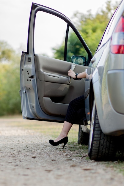 Une femme d'affaires posant près d'une voiture blanche et montrant ses jambes minces et minces près de sa voiture Une femme prospère en jupe grise posant sur des talons hauts roses