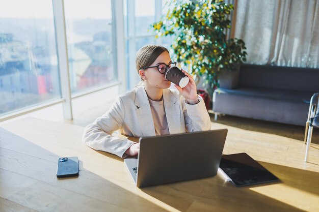 Photo une femme d'affaires positive dans une veste est assise à une table travaillant sur un ordinateur portable dans un bureau lumineux une jeune femme européenne est assise à une table travail indépendant et à distance mode de vie des femmes modernes