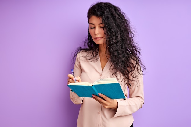 Femme d'affaires prendre des notes isolées sur le mur violet, femme brune bouclée avec ordinateur portable et stylo en mains